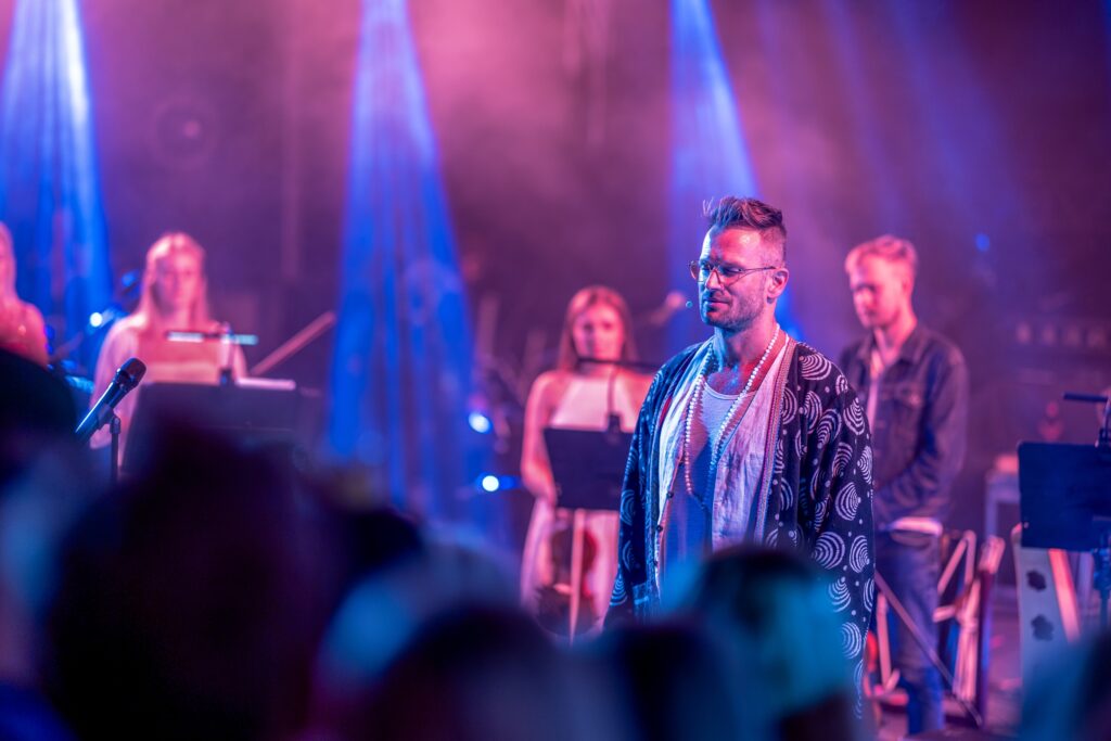 Johann Urb seisab püramiidikujulise valgusinstallatsiooni ees Meelte Videviku festivalil. Ta hoiab mikrofoni ja suhtleb kohalviibijatega, ümberringi õhtune loodus ja sumedus.